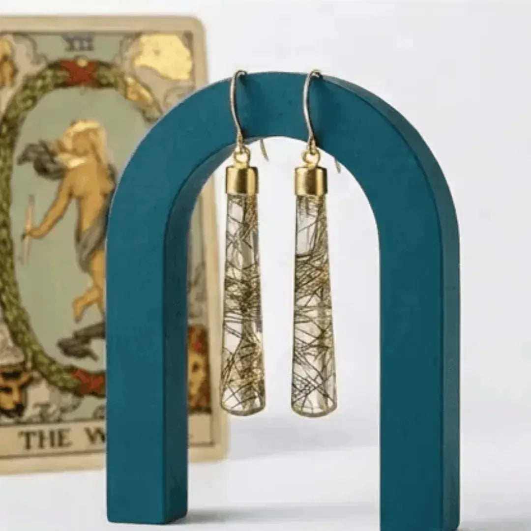Pair of gold and clear cylindrical earrings on a teal arch with a tarot card in the background.