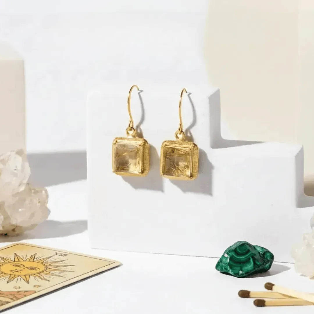 Gold square earrings on a white stand with crystals and cards in the background
