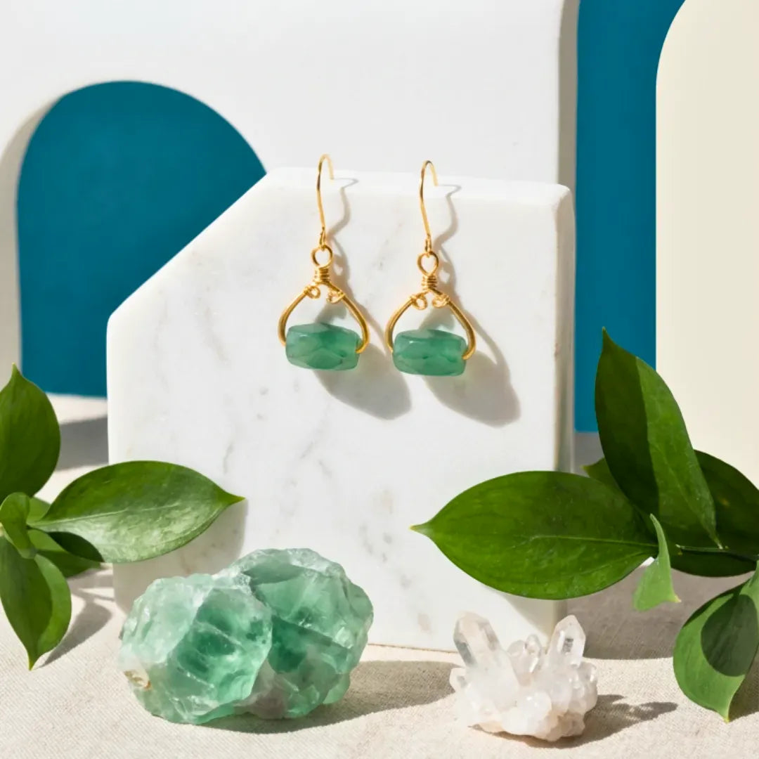 Gold earrings with green gemstones on a white stand with green leaves around.