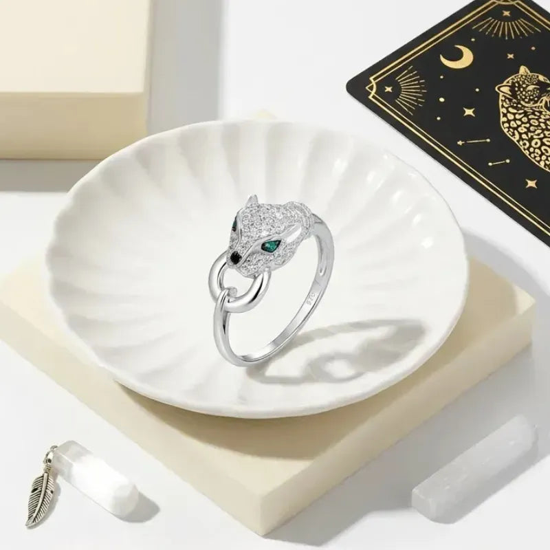 Silver panther ring with green gemstone on a white dish, with a black card featuring a panther design in the background.