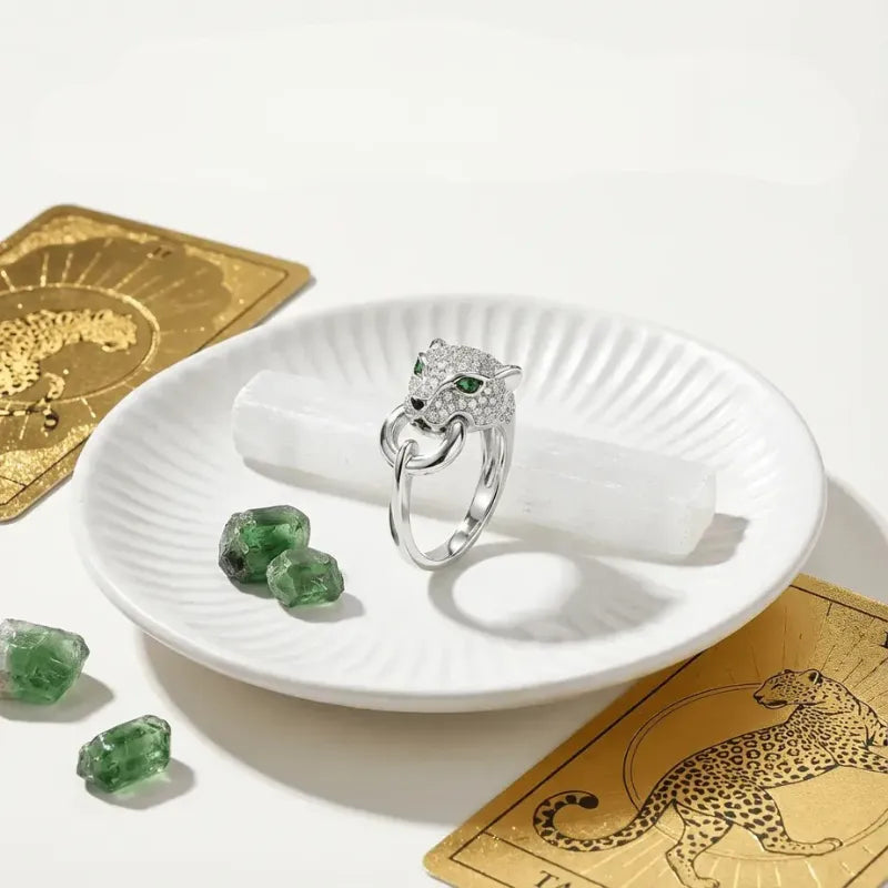 Silver ring with green gemstones on a white dish, surrounded by green crystals and gold cards with leopard designs.