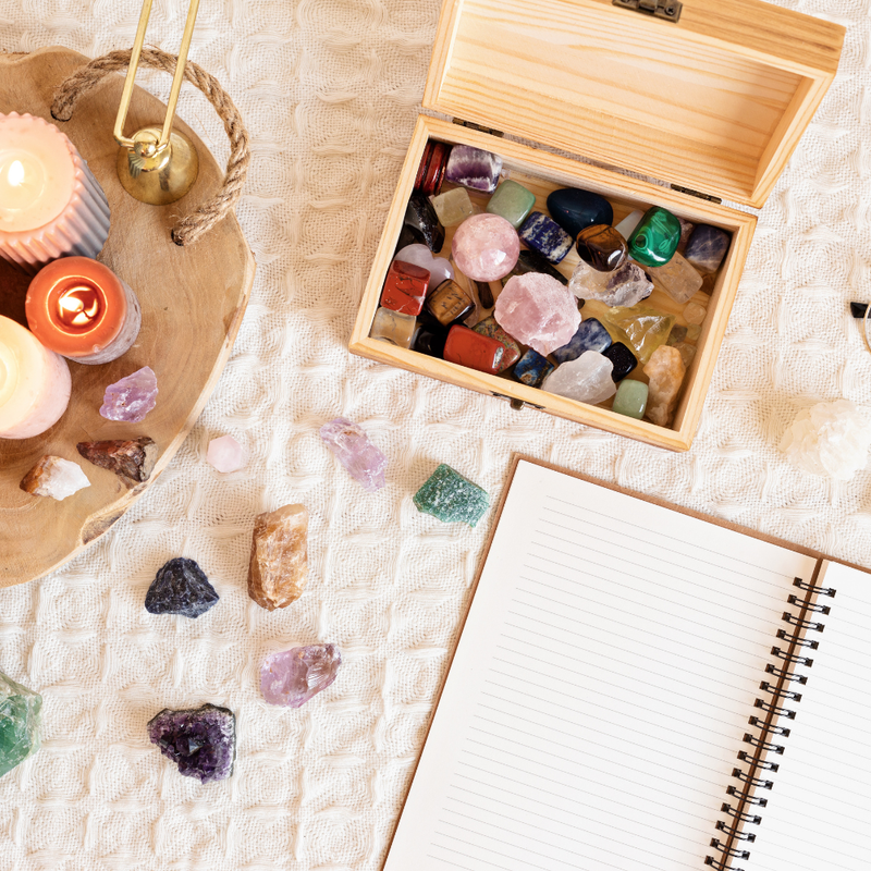 Box of colorful crystals on a textured surface with candles and a notebook.