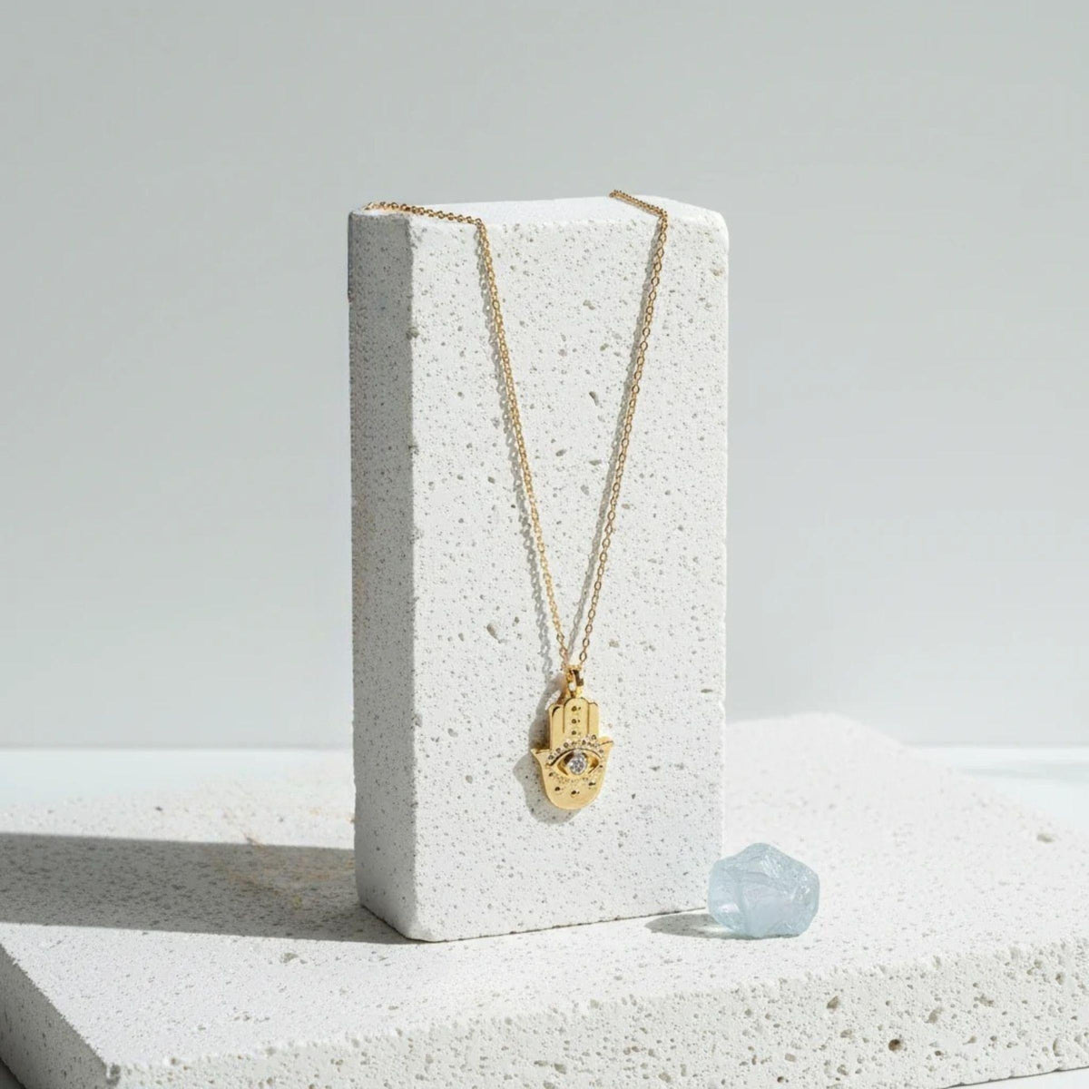 Large gold plated hamsa hand necklace hanging on a white stone display