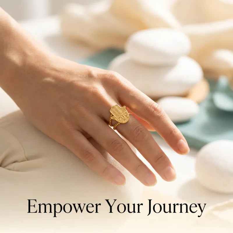 Hand wearing a gold ring with a hamsa design, text 'Empower Your Journey' on a blurred background.