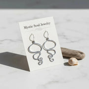 Silver earrings on a 'Mystic Soul Jewelry' card with a shell and driftwood on a light background