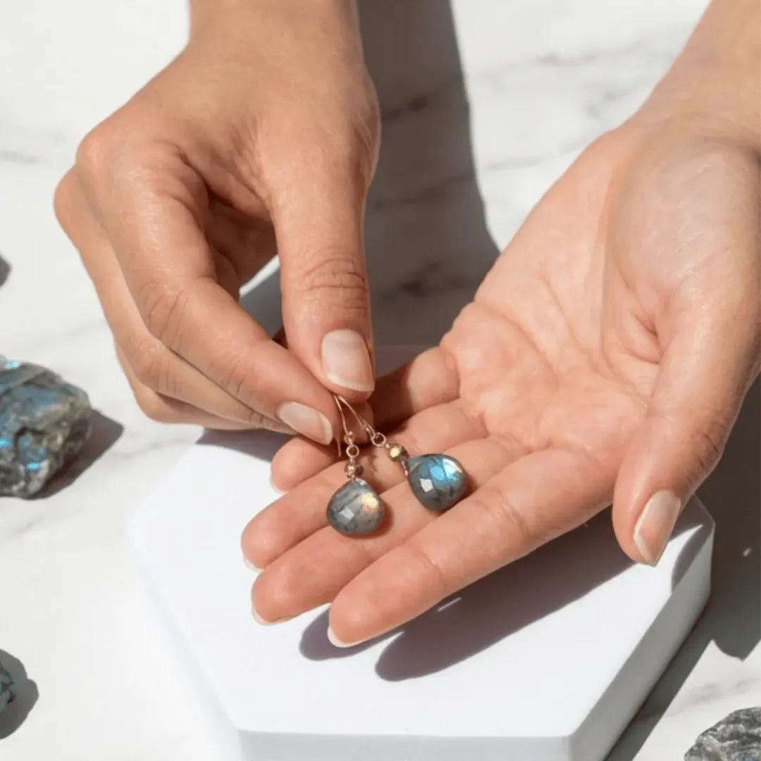 Close-up of hands holding a pair of earrings with Labradorite stones on a light background.