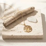Gold necklace with a circular pendant on a stone surface with driftwood and sea glass.