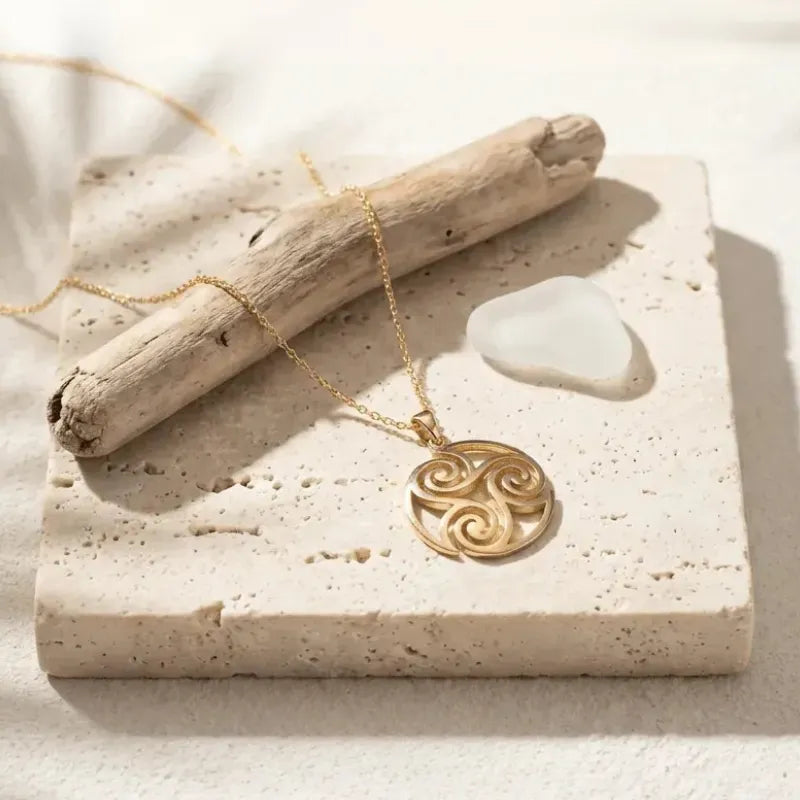 Gold necklace with a circular pendant on a stone surface with driftwood and sea glass.