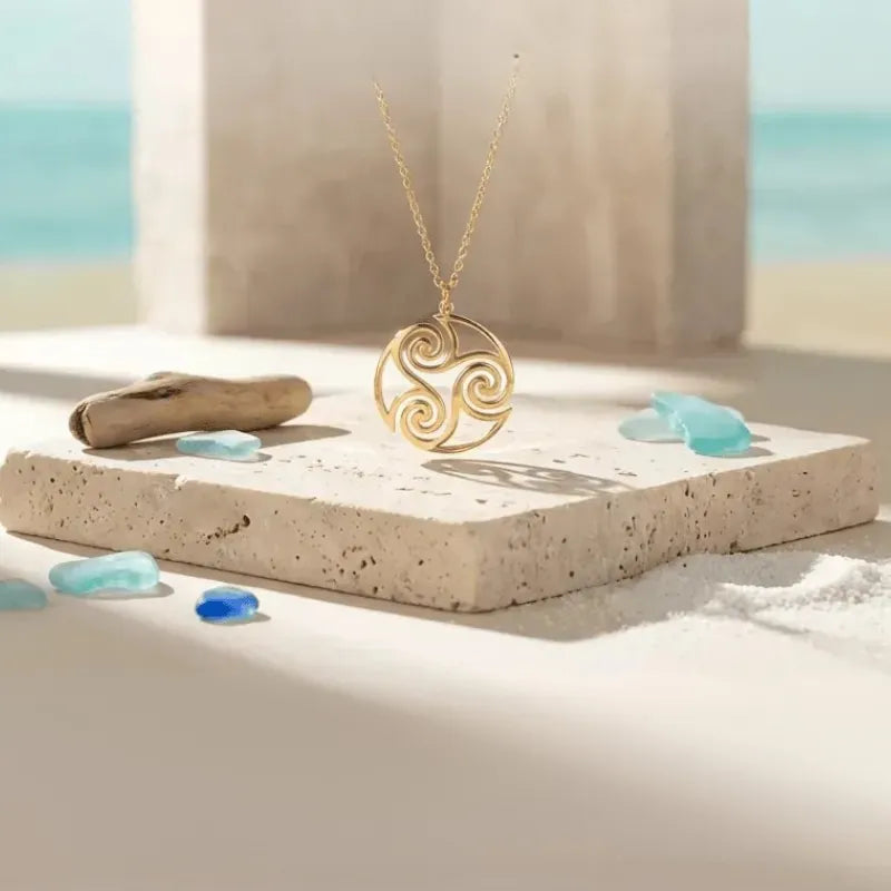 Gold necklace with a triskele pendant on a sandy surface with blue stones and ocean view