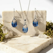 Blue teardrop jewelry set with gold chains on a marble surface with greenery.