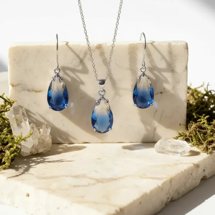 Blue teardrop jewelry set with gold chains on a marble surface with greenery.