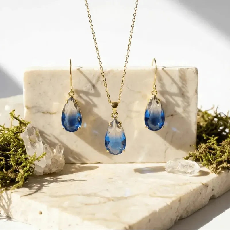 Jewelry set with blue teardrop pendants on a marble surface with greenery.