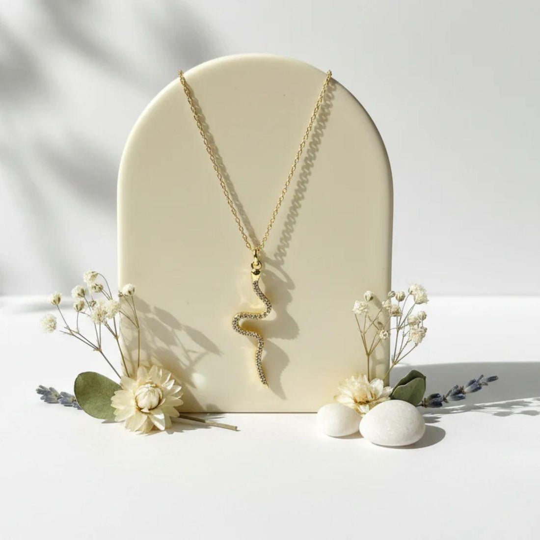 A gold snake pendant necklace displayed on a cream-colored arched stand, styled with dried flowers