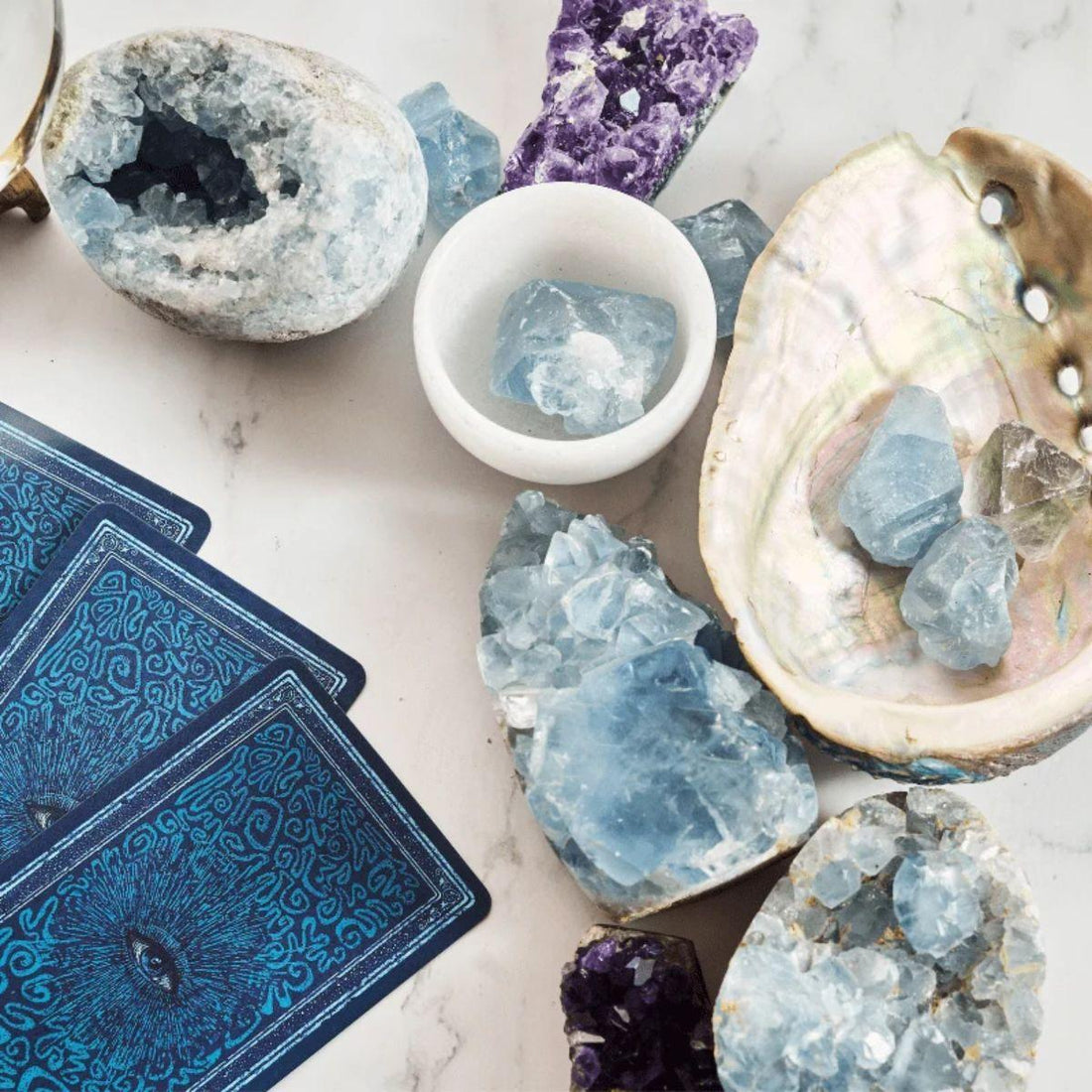 A styled flat-lay of blue crystal geodes, a white bowl of blue crystals, an open abalone shell with crystals