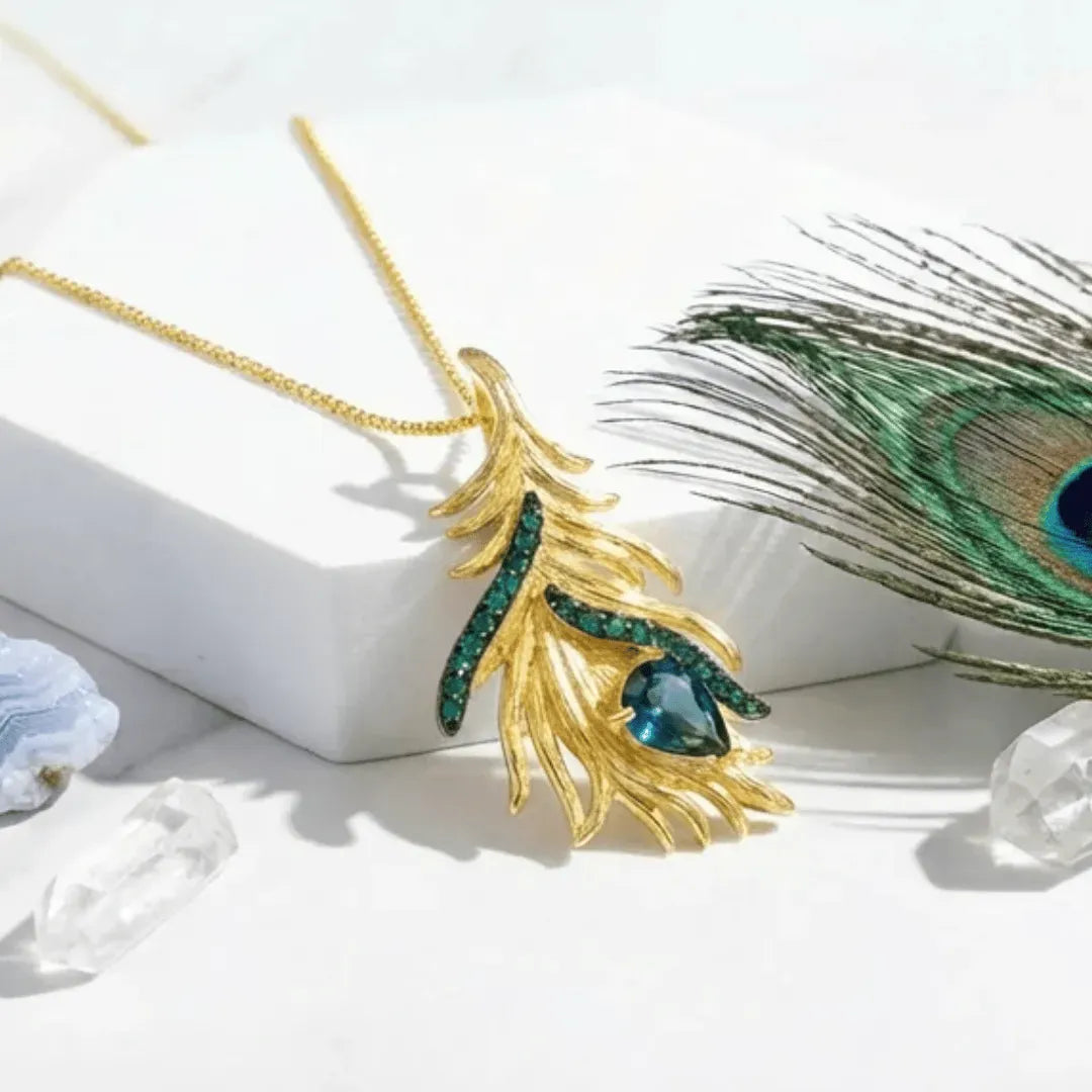 Gold feather-shaped pendant with green gemstones on a white background