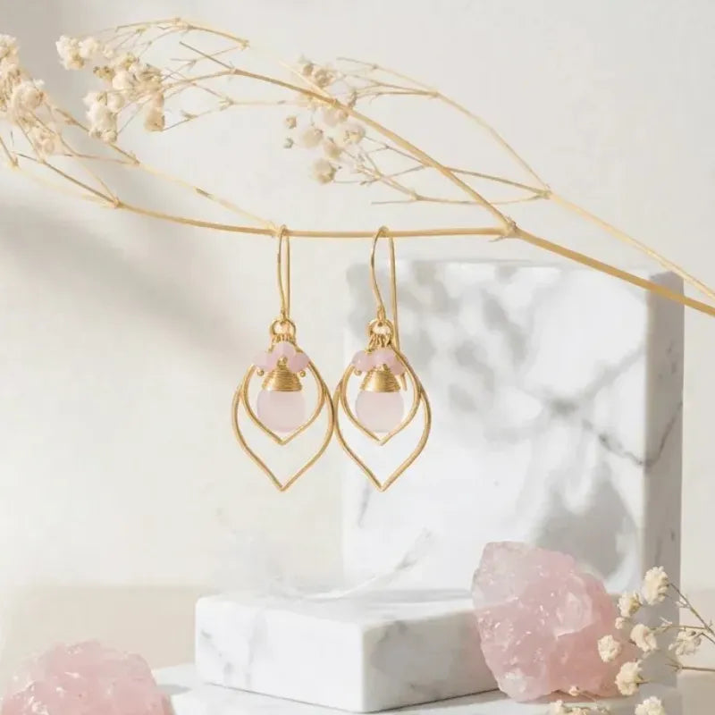 Gold earrings with pink gemstones on a marble stand with dried flowers.