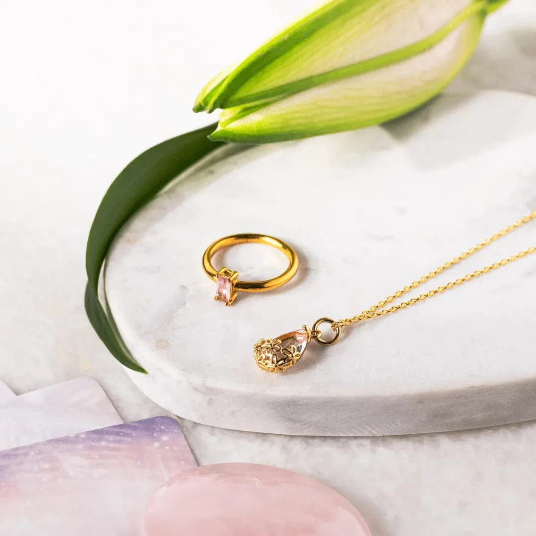 Gold ring and necklace with pink gemstones on a marble surface with green leaves.