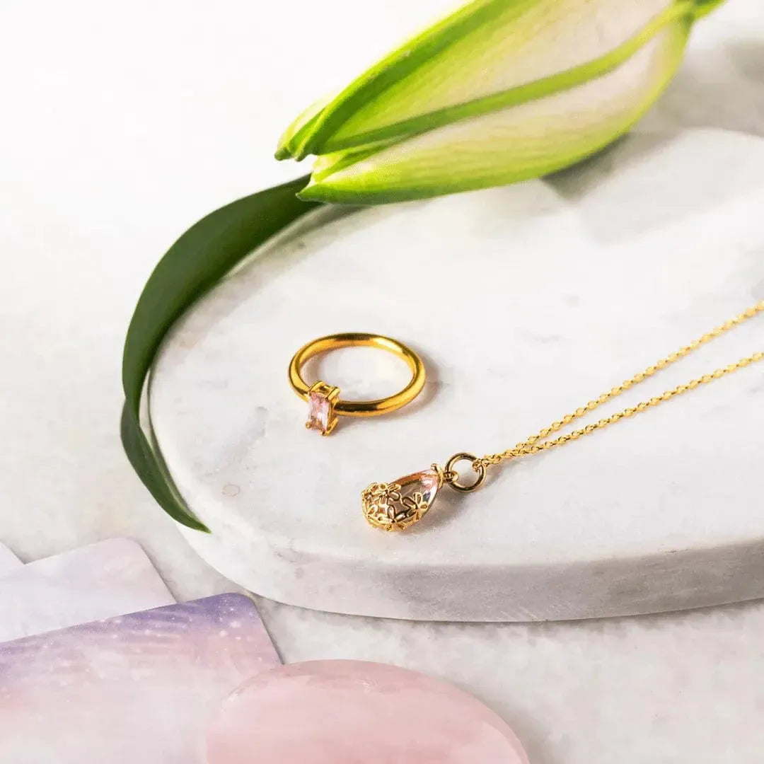 Gold ring and necklace with pink gemstones on a marble surface with green leaves.