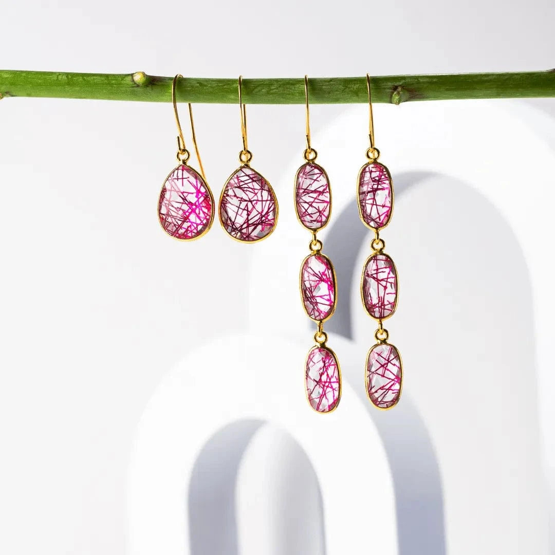 Pink teardrop earrings with gold hooks hanging from a branch on a white background