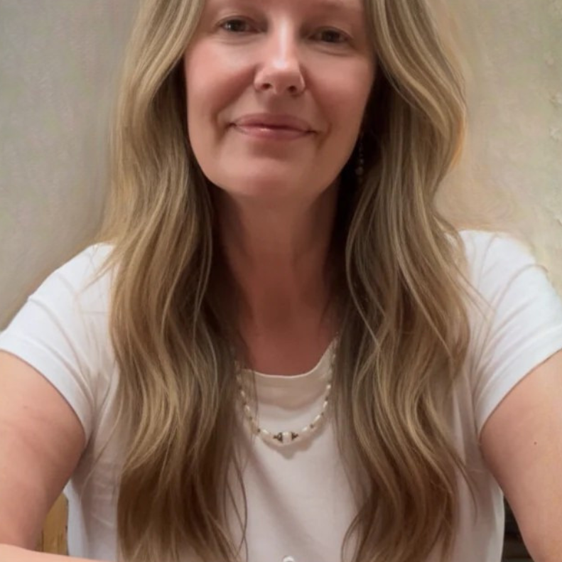 Woman with long blonde hair wearing a white shirt and pearl necklace against a neutral background