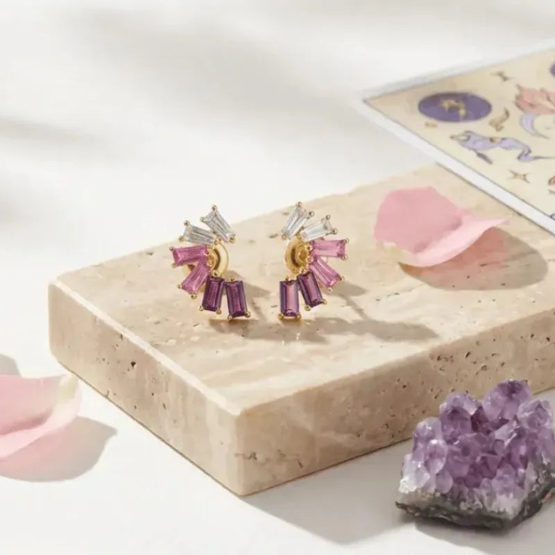 Pair of gold and purple earrings on a stone surface with pink petals and a crystal.