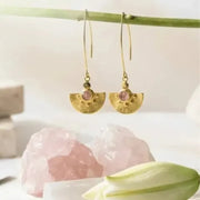 Gold earrings with pink stones on a white surface with a stone and leaf in the background