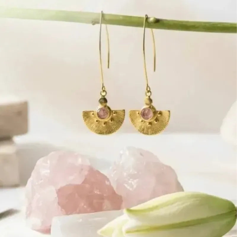 Gold earrings with pink stones on a white surface with a stone and leaf in the background