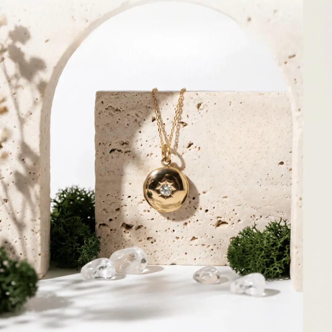 Gold necklace with a pendant on a textured stone surface with decorative stones and plants.