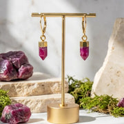 Gold earrings with pink gemstones on a gold stand, surrounded by stones and plants.