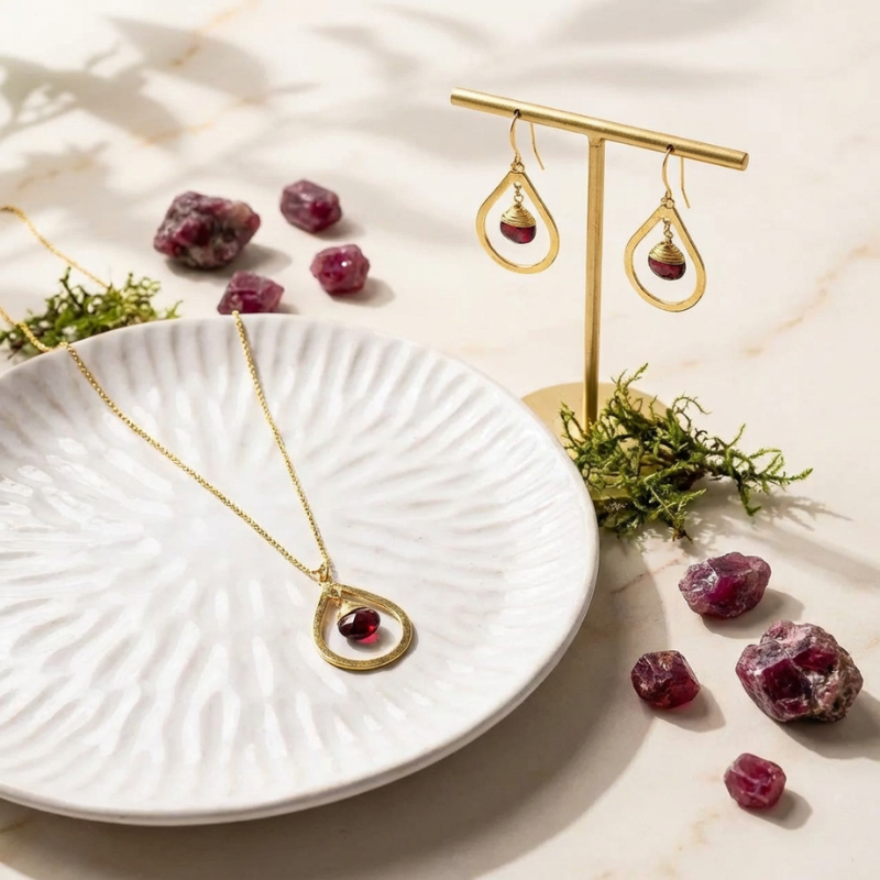 Gold jewelry set with pendant necklace and earrings on a white plate with red stones and greenery.