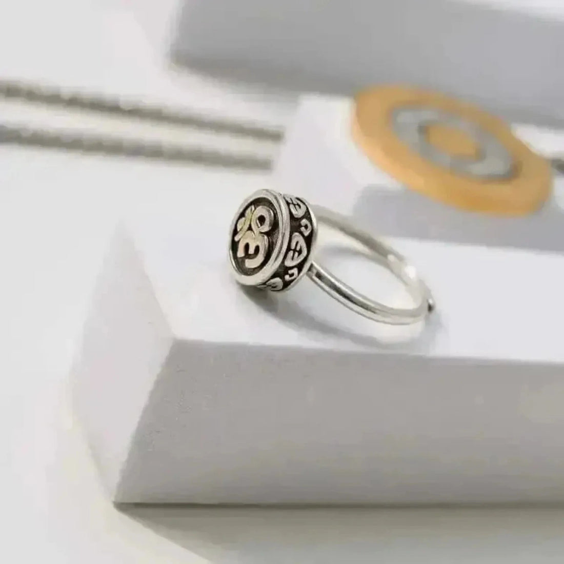 A silver ring with the Om symbol in the center, displayed on a white box with a golden chain and a round charm in the background.