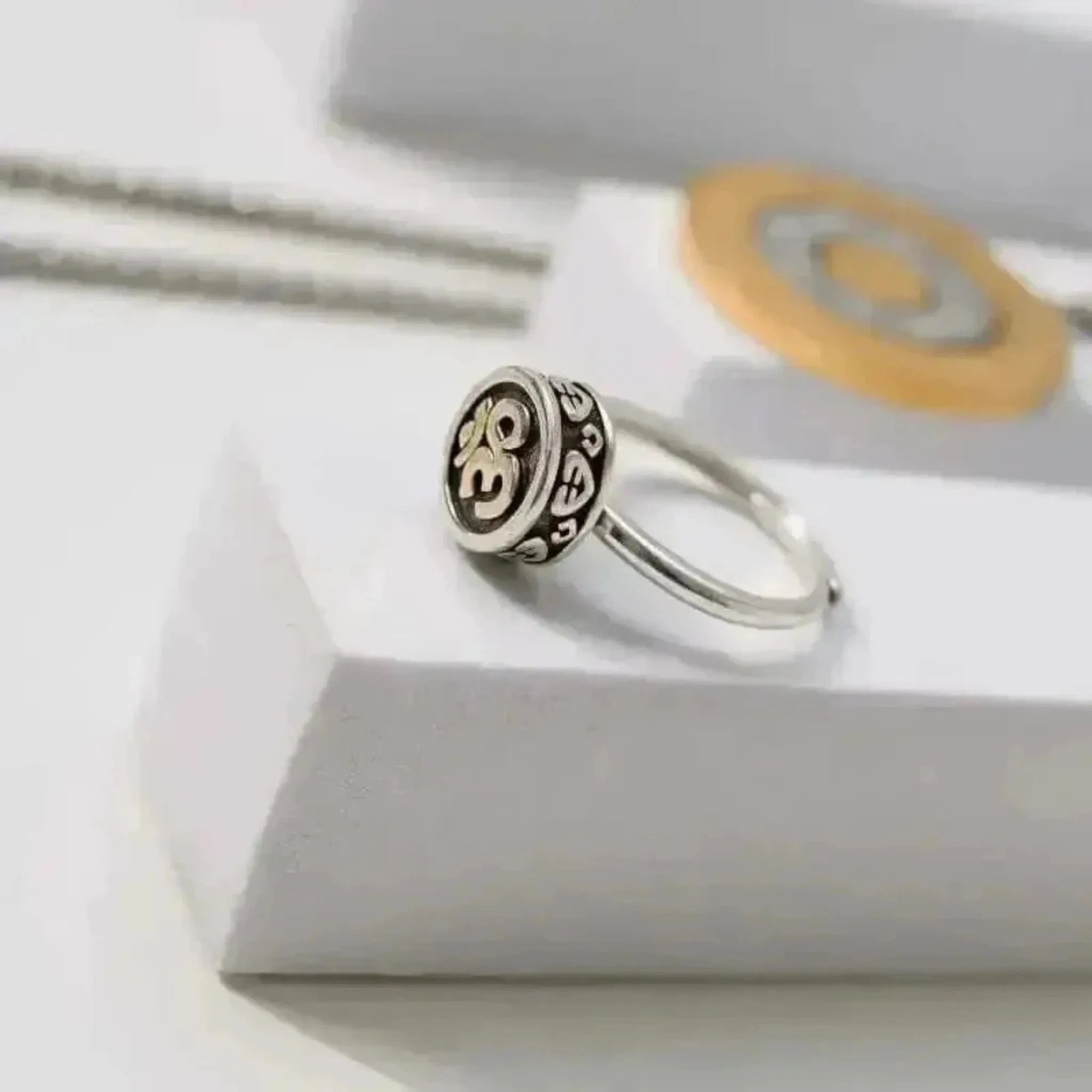 A silver ring with the Om symbol in the center, displayed on a white box with a golden chain and a round charm in the background.
