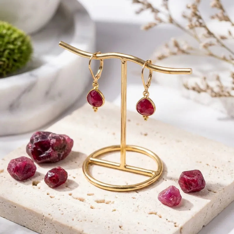 Gold earrings with red gemstones on a gold stand with scattered gemstones on a light surface.