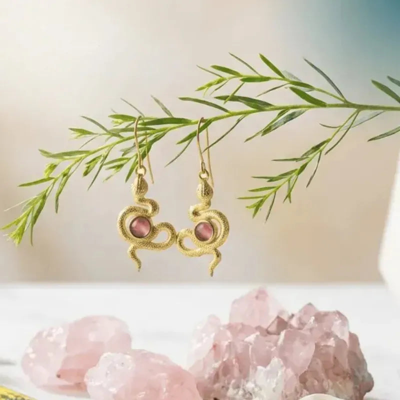 Gold earrings with pink stones on a white surface with green leaves and pink crystals in the background