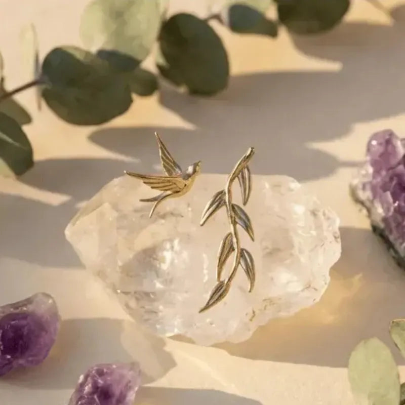 Gold bird and leaf earrings on a crystal with purple crystals and green leaves in the background