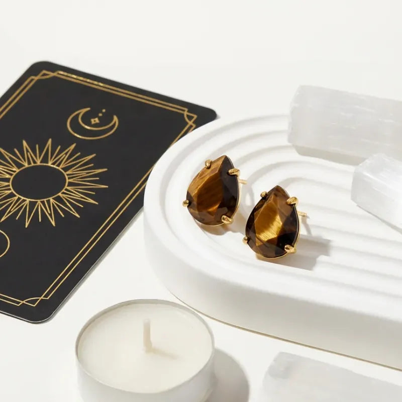 Brown gemstone earrings in a white box with a tarot card and candle on a white surface