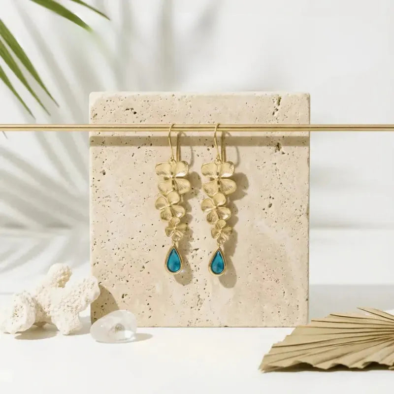 Gold earrings with blue teardrop stones on a beige stand with natural elements.