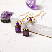 Purple beaded earrings with gold accents on a textured white surface with purple crystals.