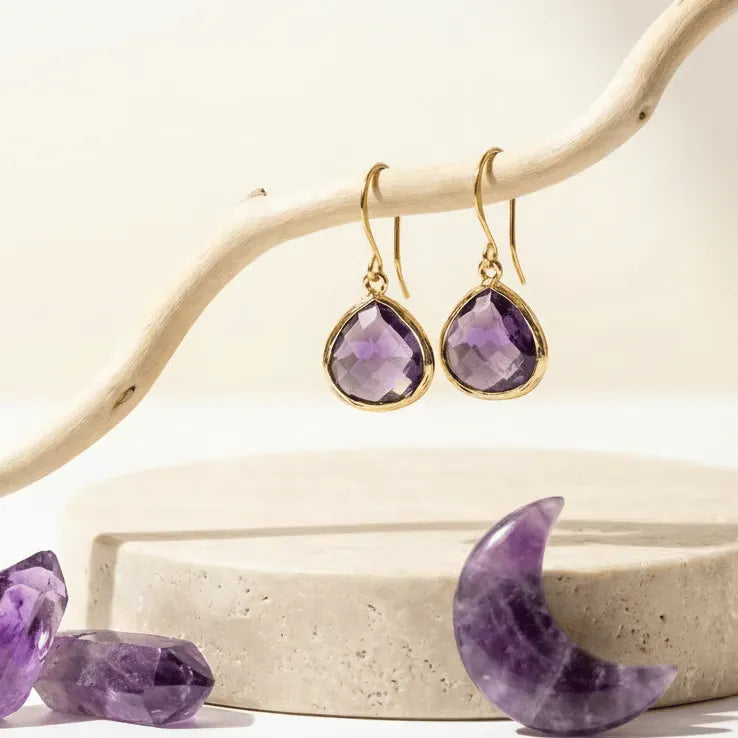 Amethyst earrings with gold hooks on a decorative stand with amethyst stones.