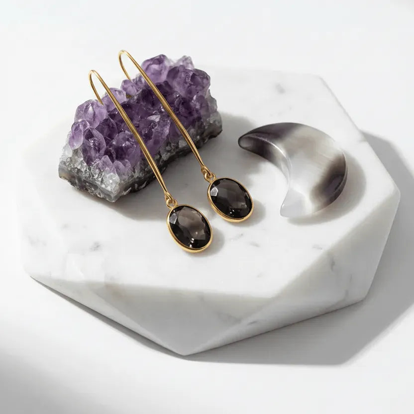 Gold earrings with smoky stones on a white marble surface with amethyst crystal.