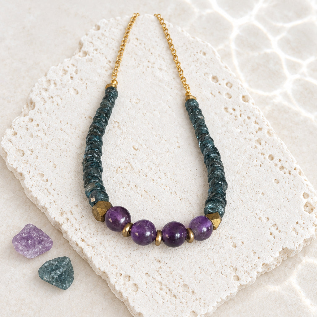 Amethyst and deep teal kyanite beaded necklace with Nepali brass accents on a gold chain, laid on white stone beside raw amethyst and kyanite