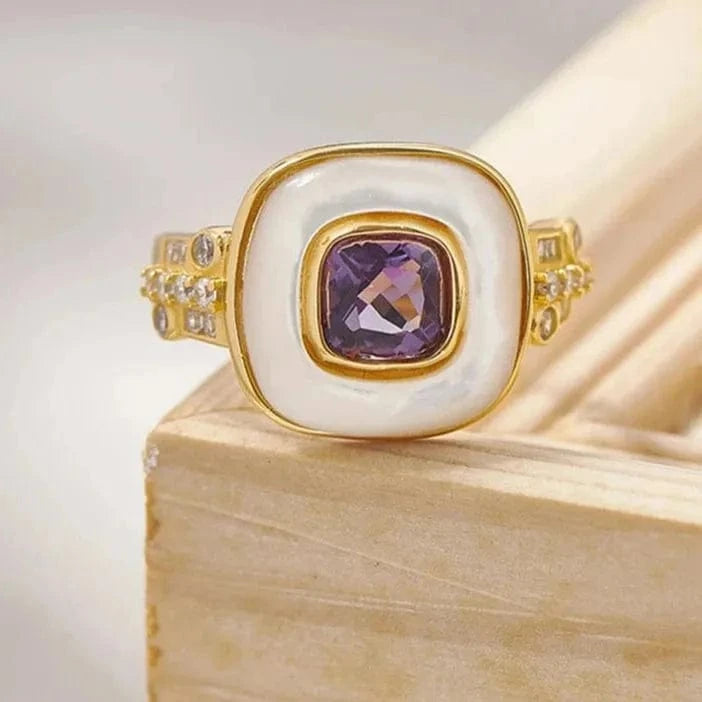 Gold ring with a purple gemstone on a wooden block, featuring the brand 'GEEZENCA'.