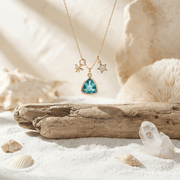 Gold necklace with a blue pendant on a sandy surface with shells and driftwood