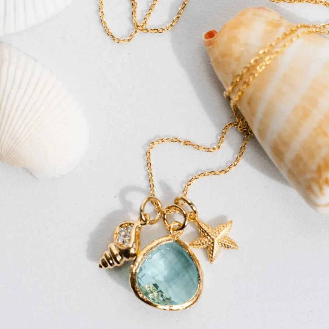 Gold necklace with a blue pendant and starfish charm on a white surface with shells.