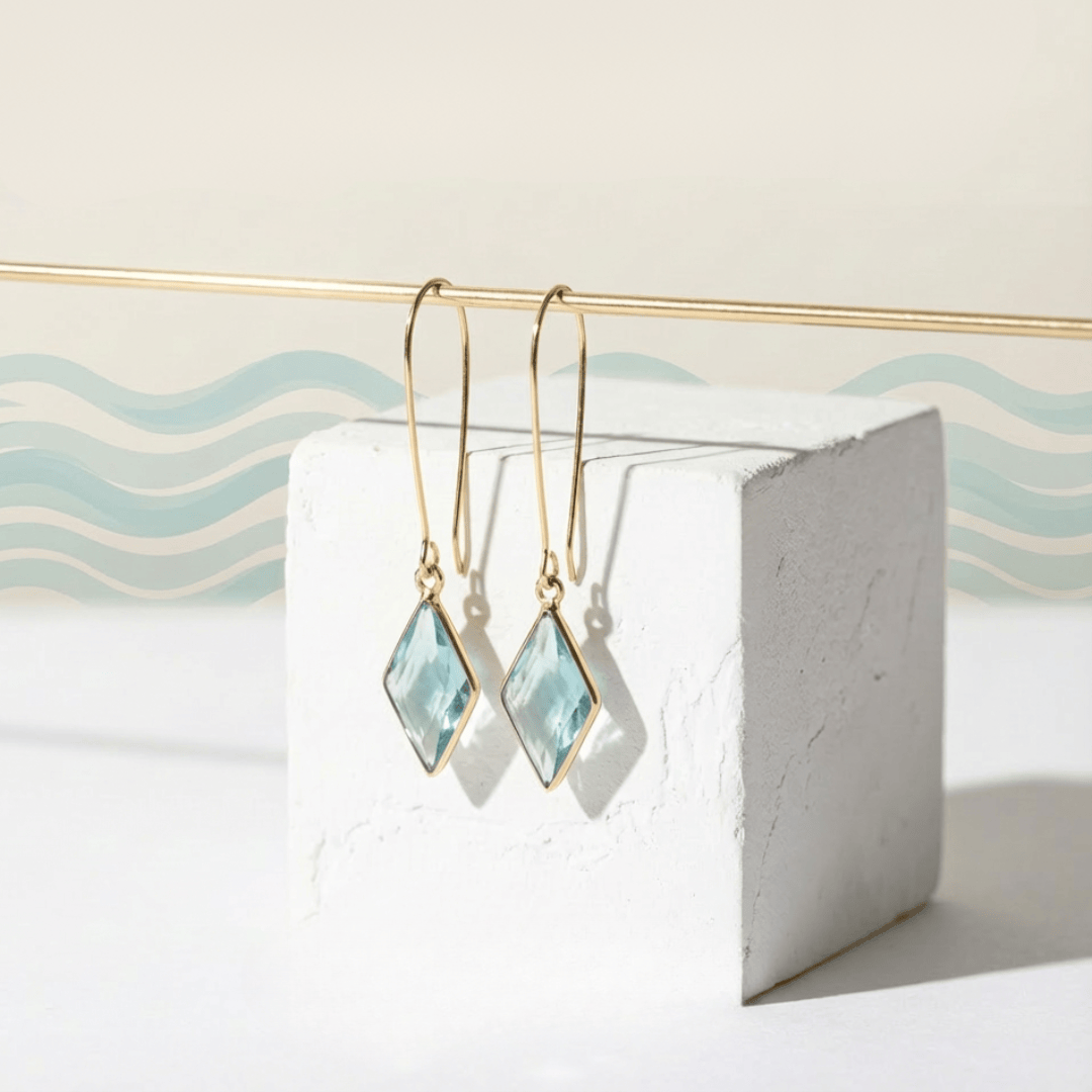 Gold earrings with light blue gemstones on a white stand with wavy blue and beige background
