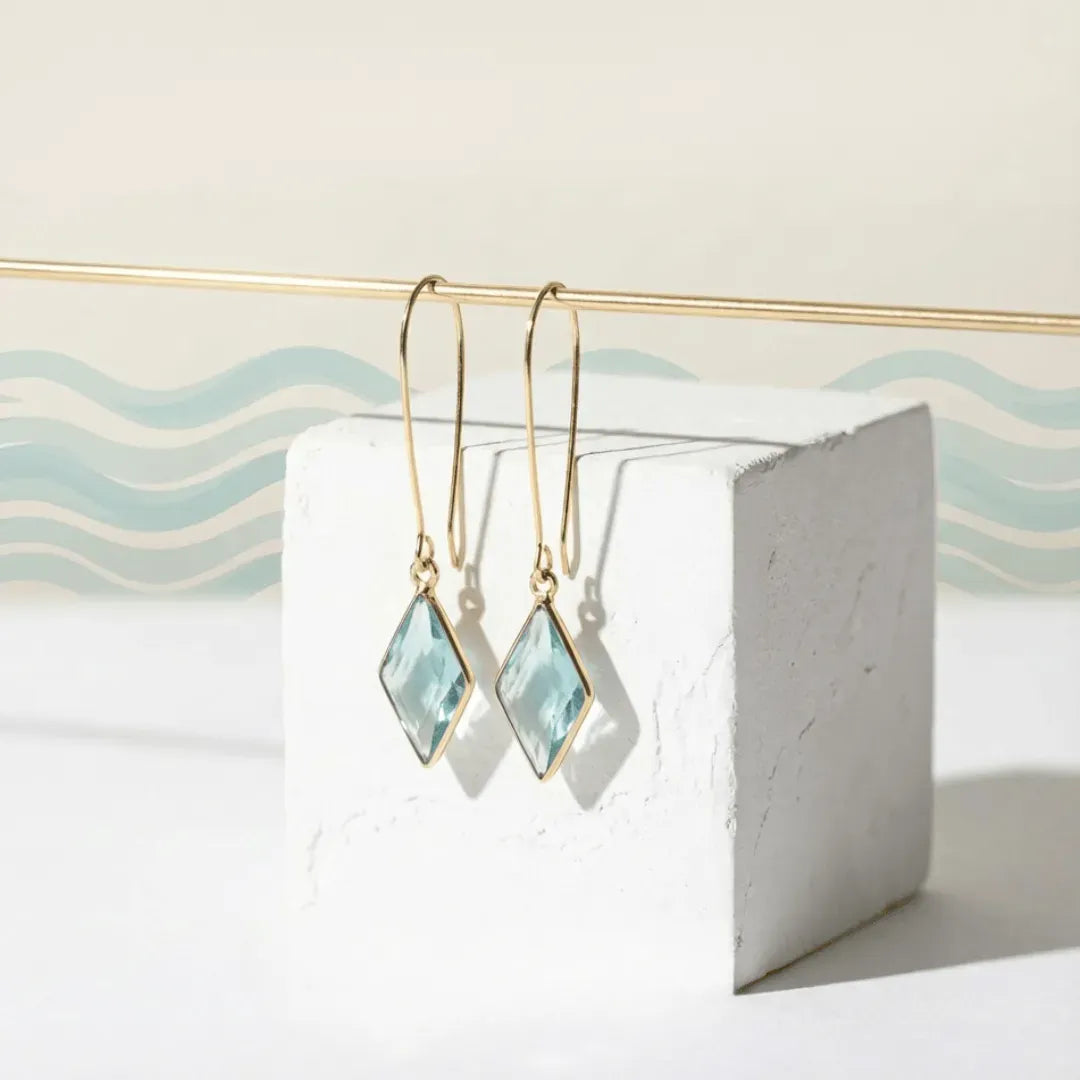 Gold earrings with light blue gemstones on a white stand with wavy blue and beige background