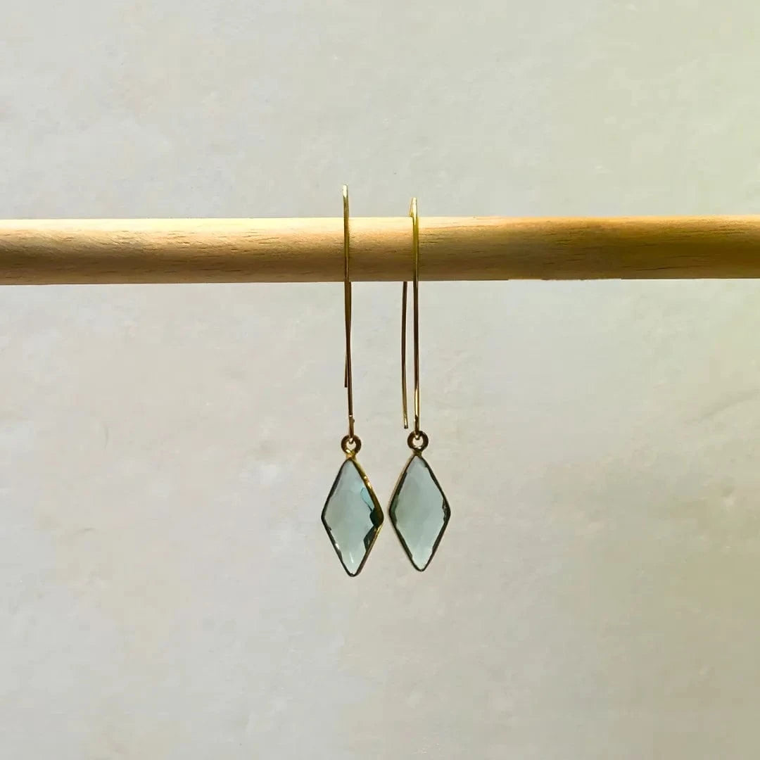 Pair of gold earrings with green teardrop-shaped stones on a wooden rod against a beige background.