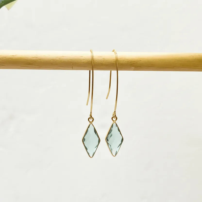 Gold earrings with green gemstones hanging on a wooden rod against a white background