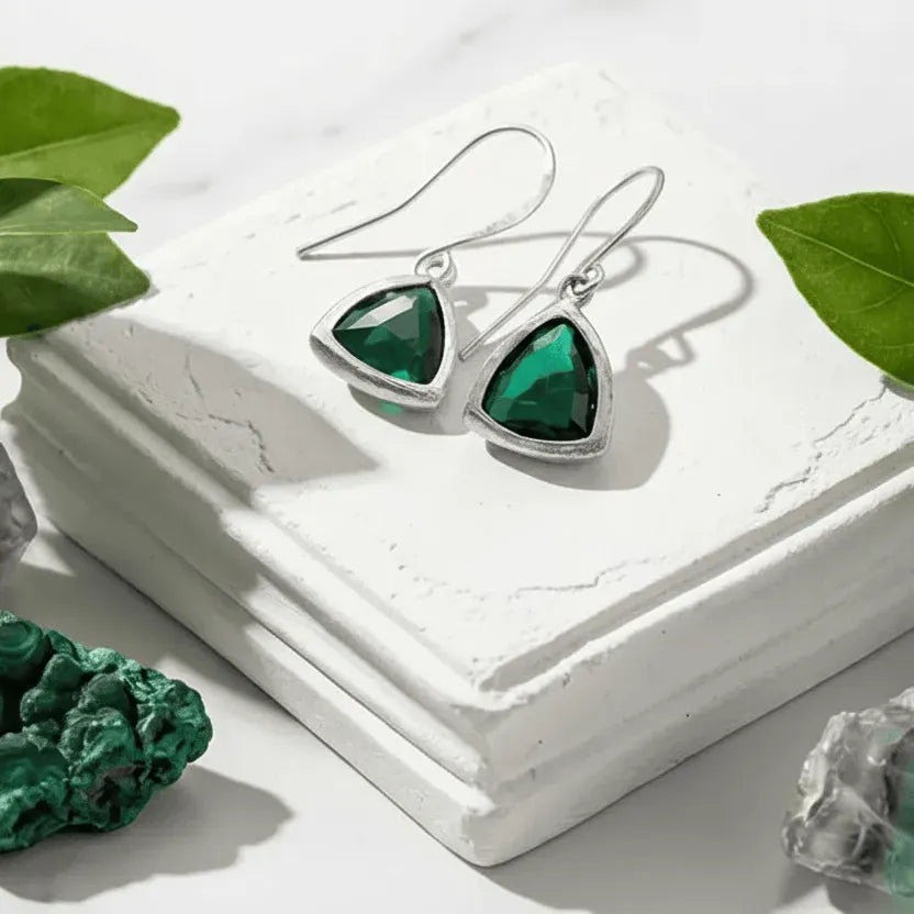 Silver earrings with green gemstones on a white surface with green leaves and crystals.