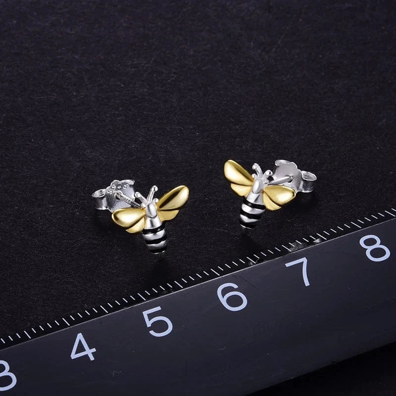 Bee-shaped earrings in gold and silver on a black background with a ruler for scale.